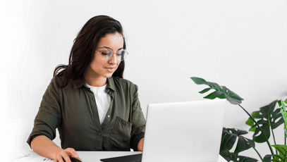 Woman working on laptop with green plants nearby.