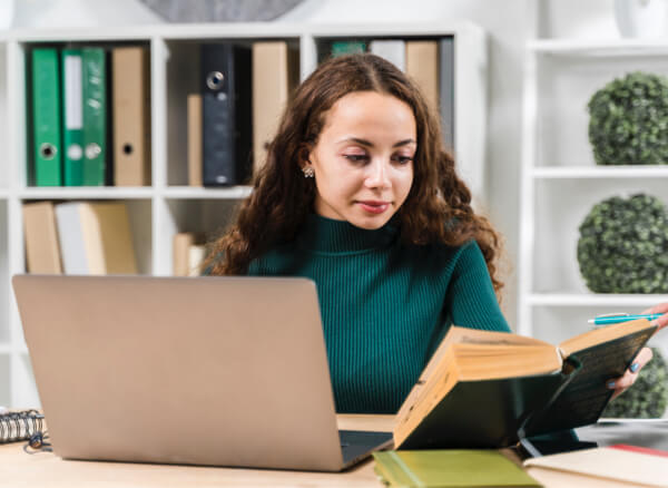 Woman reading book at office desk with laptop.